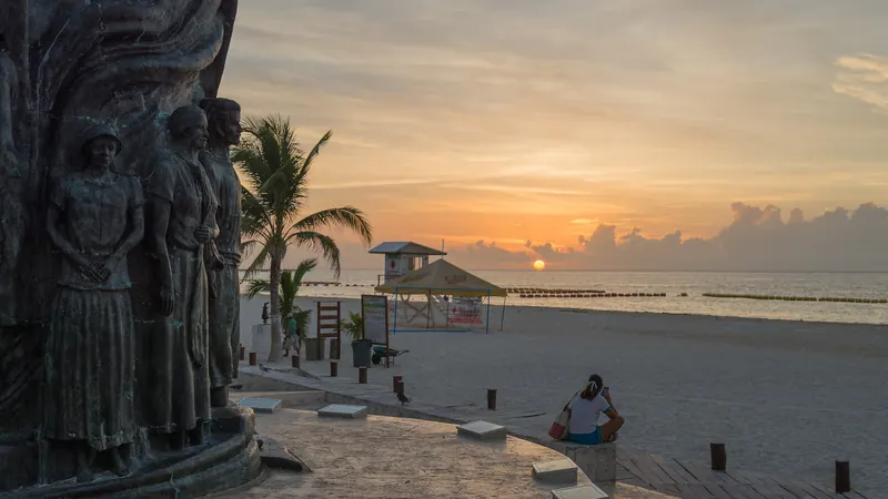 Escultura del Portal Maya en el Parque Fundadores al amanecer, con una persona sentada frente al mar en Playa del Carmen