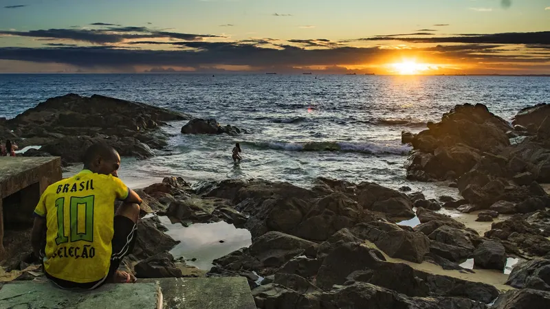 Pessoa de camisa amarela sentada nas pedras do Porto da Barra, em Salvador, com o sol se pondo sobre a Baía de Todos-os-Santos